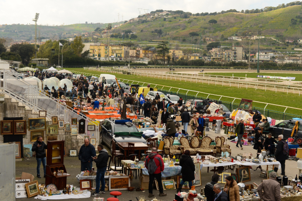 Große Gruppe von Menschen auf einem Flohmarkt mit Tischen, auf denen Gegenstände wie Fotorahmen und Stühle ausgelegt sind, Fahrzeuge in der Nähe geparkt, Geländer, Treppen, Bäume, Gebäude, Laternenpfähle, Hügel und ein bewölkter Himmel im Hintergrund.
