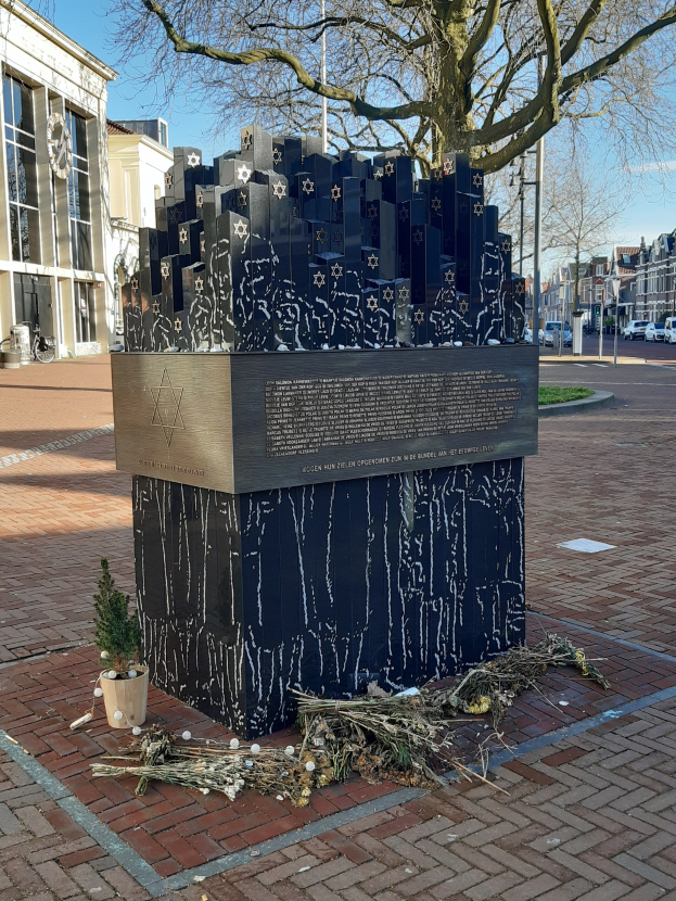 Denkmal in einem Stadtplatz für Holocaust-Opfer, mit einem Baum davor, einer Topfpflanze auf dem Boden, verstreuten trockenen Blättern und Gebäuden, Fahrzeugen, Pfählen und Bäumen im Hintergrund.