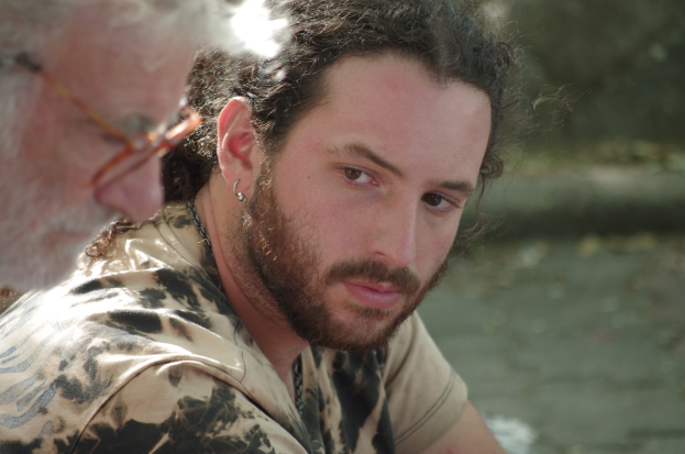 Zwei Männer sitzen nebeneinander bei einem Gewässer, einer trägt eine Brille, mit einem sanft unscharfen Hintergrund.