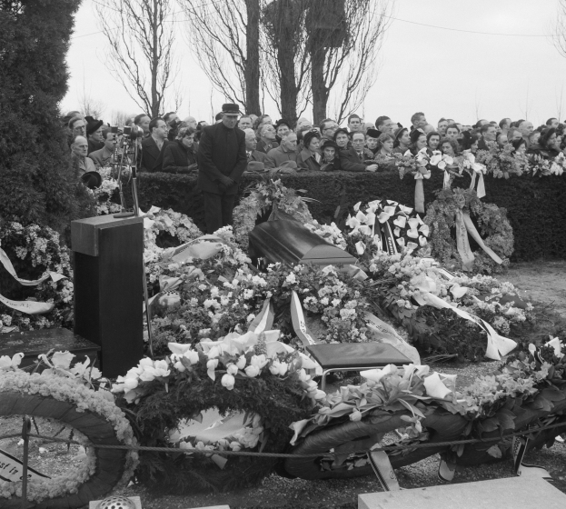Schwarze und weiße Trauerprozession mit einem Mann in einer Mütze, der auf einem Podium spricht und von Blumen, Stühlen und einer Menge umgeben ist, vor einem Hintergrund mit Bäumen und einem klaren Himmel.