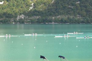 Eine Gruppe von Menschen, die auf einem Gewässer umgeben von Bäumen und einem Hügel rudern, mit Ständen, die Kleidung zeigen, am unteren Bildrand, was auf einen Ruderwettbewerb hindeutet.