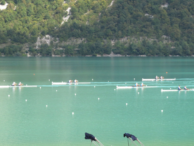 Eine Gruppe von Menschen, die auf einem Gewässer umgeben von Bäumen und einem Hügel rudern, mit Ständen, die Kleidung zeigen, am unteren Bildrand, was auf einen Ruderwettbewerb hindeutet.