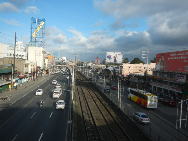 Eine belebte Stadtstraße mit Verkehr, neben einem Bahngleis, mit Gebäuden, Strommasten, Schildern, Bäumen und einem bewölkten Himmel.