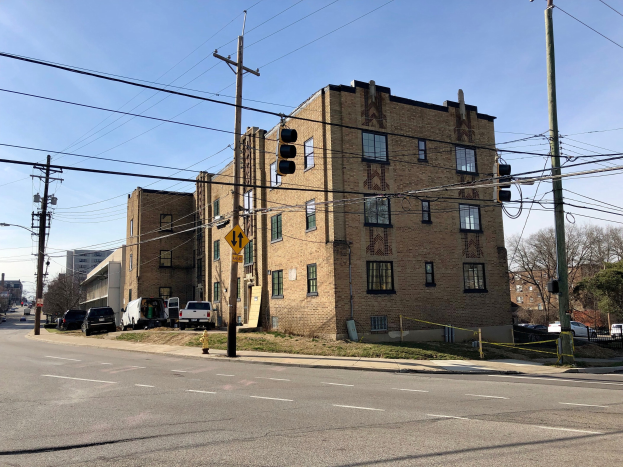Großes Backsteingebäude an einer Straßenecke mit Fahrzeugen, Ampeln, Schildern, Strommasten und Bäumen im Hintergrund.