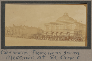 Eine Gruppe deutscher Kriegsgefangener aus Messines steht vor einem Gebäude unter einem klaren blauen Himmel, mit Text am unteren Bildrand.