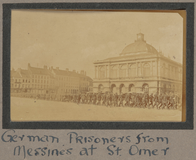 Eine Gruppe deutscher Kriegsgefangener aus Messines steht vor einem Gebäude unter einem klaren blauen Himmel, mit Text am unteren Bildrand.