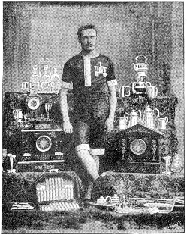 Ein Schwarz-Weiß-Foto eines Mannes in einem Fußballtrikot, der vor einem Tisch mit Pokale und Trophäen steht, mit der Beschriftung "1909-1910 WM-Sieger" unten.