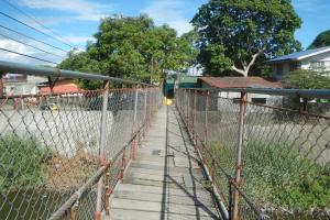 Eine Hängebrücke überspannt einen Fluss, flankiert von Zäunen, mit üppiger Vegetation, Häusern und einem klaren blauen Himmel im Hintergrund.