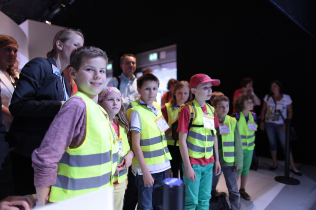 Eine Gruppe von Kindern in Sicherheitwesten und Namensschildern steht auf einem Boden, mit einem Jungen auf der linken Seite, der l√§chelt, einem Pfahl auf der rechten Seite und ein paar Menschen im Hintergrund.