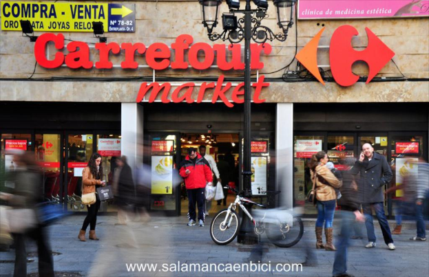 Ein belebter Einkaufsmarkt mit Menschen vor einem Einkaufszentrum, ein schwarzer Pfahl mit Lichtern, ein Fahrrad neben dem Pfahl und ein Richtungsschild an der Wand.