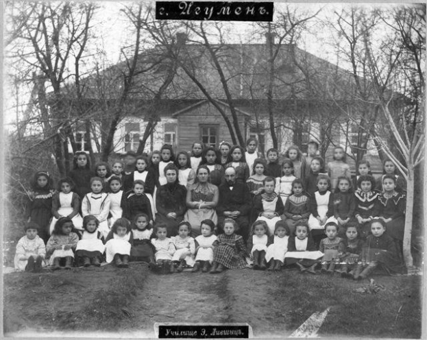 Schwarzes und weißes Foto einer Gruppe von Kindern, die vor einem Gebäude mit Fenstern posieren, umgeben von Bäumen und einem klaren Himmel, mit Text unten, der 'Jugoslawien, die erste Schule der Welt' liest.
