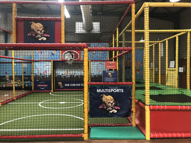 Großes Indoor-Trampolin-Park mit einem Basketballfeld in der Mitte, umgeben von einem Netz, mit einem Basketballkorb, Deckenleuchten und Wandbannern.