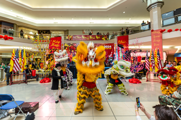 Ein lebendiges chinesisches Neujahrsfest in einem Einkaufszentrum mit tanzenden Menschen in Kostümen, anderen auf Stühlen sitzend mit Mobiltelefonen und verschiedenen Dekorationen, Flaggen und Bannern im Hintergrund.