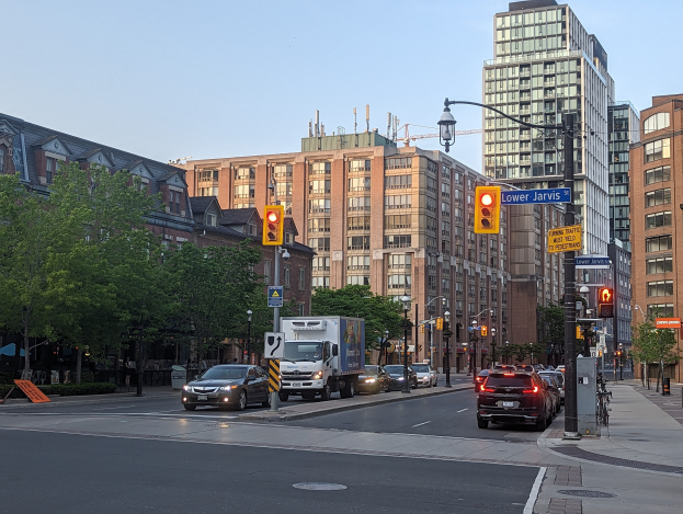Eine belebte Stadtstraße mit verschiedenen Fahrzeugen, darunter ein Lkw, ein Auto und ein SUV, unter Verkehrsampeln und Laternen, gesäumt von Gebäuden mit Fenstern und Bäumen, vor einem Himmel als Hintergrund.