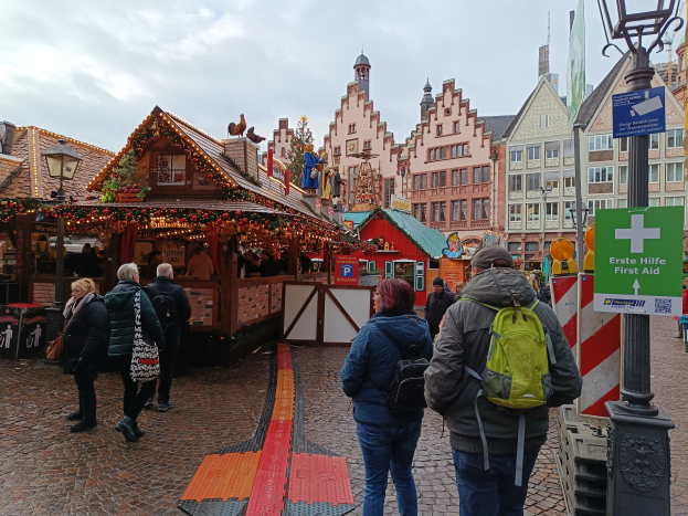 Eine Gruppe von Menschen geht eine Kopfsteinpflasterstraße in Nürnberg, Deutschland, entlang, neben einem Weihnachtsmarkt, mit Laternen, Texttafeln und Gebäuden mit Fenstern im Hintergrund unter einem bewölkten Himmel.