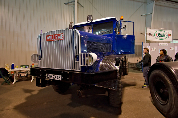 Ein Lkw steht in der Mitte des Bildes mit ein paar Menschen, die rechts daneben stehen. Im Hintergrund ist eine Wand mit einem Tisch davor, auf dem ein Krug, eine Tasse und Abdeckungen stehen, umgeben von Stühlen. Rechts daneben ist ein weiteres Fahrzeug mit einem sichtbaren Reifen.