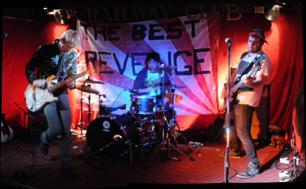 Zwei Personen vor einem Mikrofon stehend, die Gitarren spielen, während ein Mann mit Brille hinter ihnen Schlagzeug spielt, vor einem Hintergrund mit hängenden Flexi-Schallplatten und CDs.