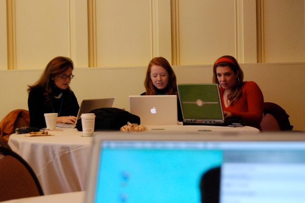Drei Frauen sitzen um einen runden Tisch und arbeiten an einem Laptop, mit einer Wand im Hintergrund.