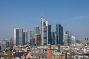 Die Skyline von Frankfurt wird von einem Hügel aus betrachtet, mit einem prominenten Wolkenkratzer in der Mitte, umgeben von anderen Gebäuden und Bäumen, unter einem klaren Himmel.