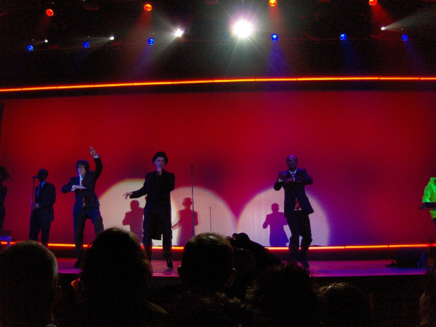 Ein Mann in einem schwarzen Kleid hält ein Mikrofon auf der Bühne, mit drei tanzenden Personen neben ihm, einem Kameramann vorne, einem Publikum, das zusieht, und einem Pianisten in einem grünen Jackett auf der rechten Seite, alles unter fokussiertem Bühnenlicht.