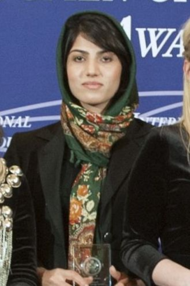 Malala Yousafzai, eine Nobelpreisträgerin für den Frieden, steht in der Mitte und trägt ein schwarzes Kleid und einen Schal, hält eine Auszeichnung und wird von zwei Frauen in schwarzen Kleidern flankiert, mit einem blauen Banner im Hintergrund.