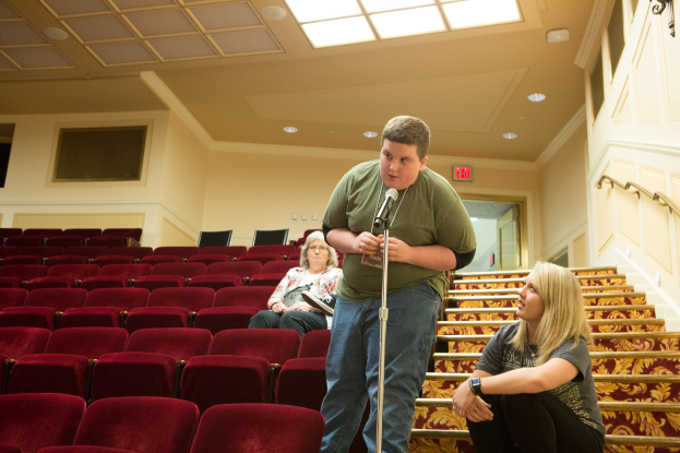 Ein Mann in einem grünen T-Shirt und blauen Jeans steht vor einem Publikum in einem Theater, hält ein Mikrofon in der Hand, zwei Frauen sitzen auf der Treppe hinter ihm, rote Stühle im Hintergrund, ein Geländer auf der rechten Seite und Lampen auf dem Dach.