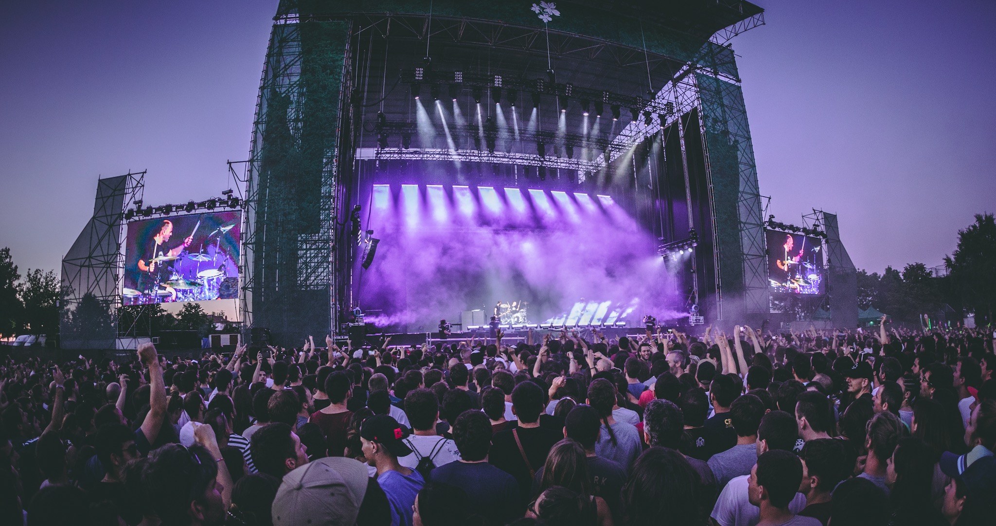 Eine große Menschenmenge bei einem Musikfestival schaut auf einer beleuchteten Bühne mit Instrumenten und Bildschirmen, vor einem Gebäude, Bäumen und einem blauen Himmel.
