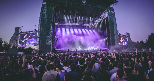 Eine große Menschenmenge bei einem Musikfestival schaut auf einer beleuchteten Bühne mit Instrumenten und Bildschirmen, vor einem Gebäude, Bäumen und einem blauen Himmel.