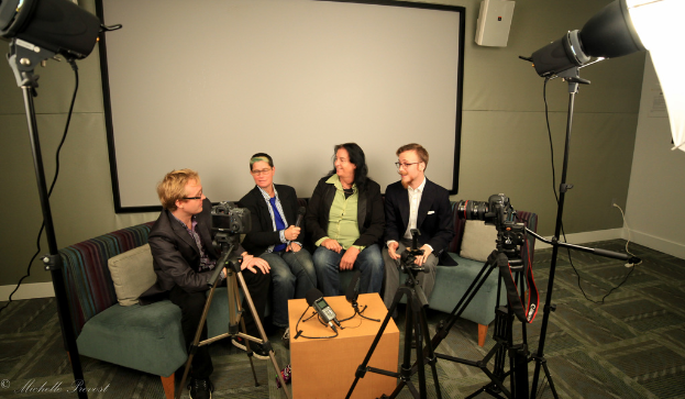 Eine Gruppe von drei Personen sitzt auf einem Sofa und gibt ein Interview, wobei eine Person in einem schwarzen Mantel und Hose links, eine Person in einem Mantel und blauer Krawatte mit einem Mikrofon in der Mitte und eine strahlende Person rechts zu sehen ist. Ein Mikrofonrekorder befindet sich auf dem Tisch in der Mitte, Kameras sind auf der rechten und linken Seite und große Scheinwerfer sind vorhanden, mit einem Projektionsschirm im Hintergrund.