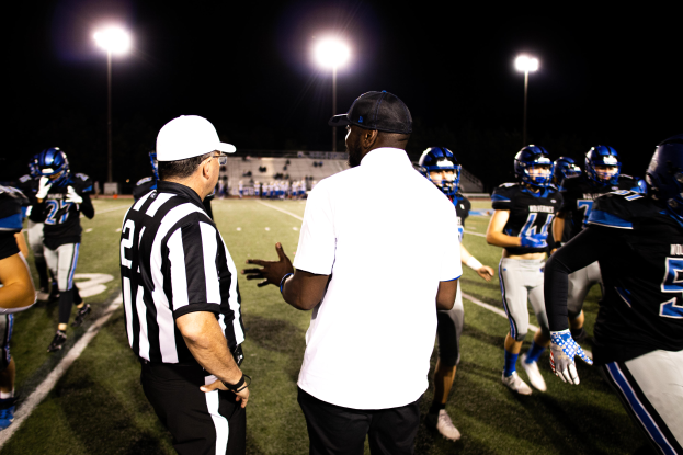 Ein Fußballtrainer in einer Mütze spricht mit seinem Team auf einem Feld bei Nacht, mit Spielern in Helmen, Laternenmasten und einem dunklen Himmel im Hintergrund.