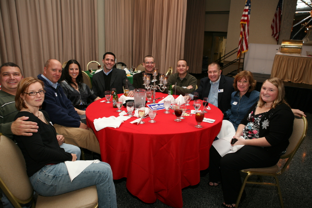 Eine Gruppe strahlender Menschen sitzt an einem Tisch mit Gläsern, Papieren, Weinflaschen und einer Flagge auf der rechten Seite und Vorhängen an der Wand.