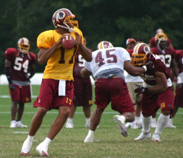 Eine Gruppe von Fußballspielern in Trikots, Helmen und weißen Schuhen auf einem Rasenfeld mit Bäumen im Hintergrund.
