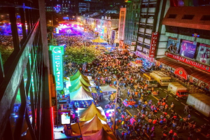 Eine pulsierende Karnevalsszene mit einer großen Menschenmenge auf der Straße, umgeben von Zelten, Geschäften, Gebäuden und Bannern, mit zahlreichen Lichtern im Hintergrund.
