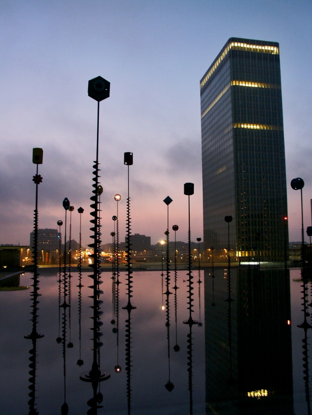 Mehrere Pfähle und Gebäude mit Lichtern unter einem bewölkten Himmel, mit ihren Spiegelungen.