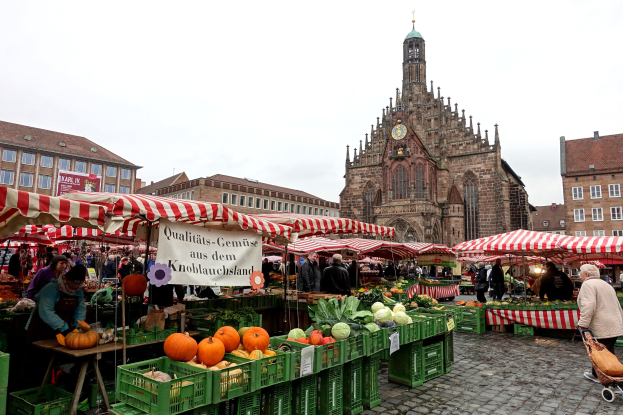 Ein belebter Markt in Nürnberg, Deutschland, mit Obst und Gemüse, Menschen mit Taschen, Zelten, Gebäuden mit Fenstern, einem Uhrenturm und einem sichtbaren Himmel.