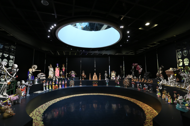 Ein großer Raum voller verschiedener Spielzeuge, darunter Skelette, die auf dem Boden angeordnet sind, mit Totenschädeln an den Wänden und Lichtern an der Decke, die eine festliche Atmosphäre für das Día de los Muertos schaffen.