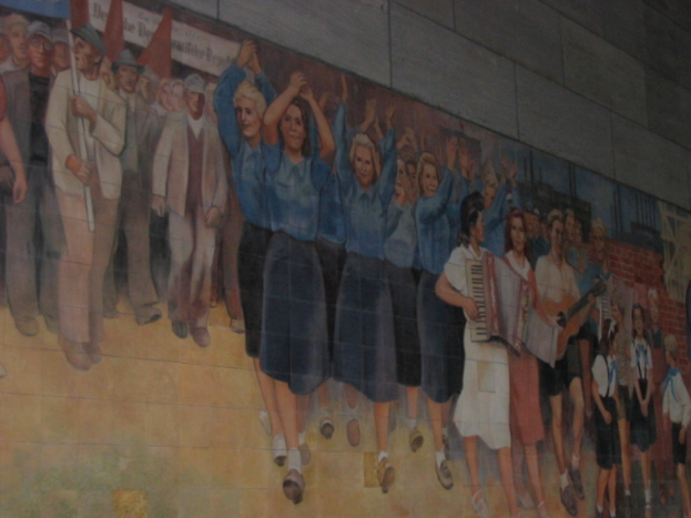 Eine Gruppe von Menschen mit blauen Mädchen in der Mitte, die klatschen, Musiker auf der rechten Seite, Kinder, die in der rechten Ecke spielen, und Individuals mit roten Fahnen auf der linken Seite.