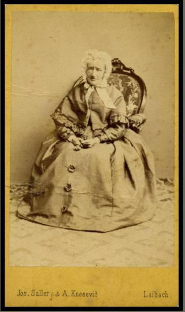 Ein altes Foto einer Frau mit sanftem Gesichtsausdruck, die ein helles Kleid und einen gemusterten Schal trägt und auf einem Stuhl sitzt, mit ihren Händen auf dem Schoß ruhend und ihrem Haar zu einem ordentlichen Knoten gebunden, in einem Photo Rahmen.