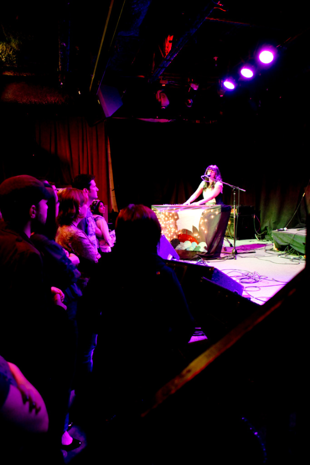 Eine Gruppe von Menschen auf der linken Seite, eine Bühne auf der rechten Seite mit Kabeln, ein Mikrofon, Frauen, ein Tisch und verschiedene Gegenstände, mit Scheinwerfern und Eisenrahmen oben und einem Vorhang im Hintergrund.