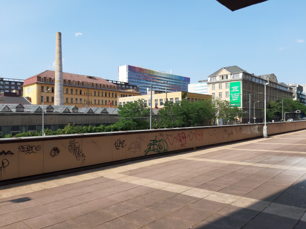 Stadtstraßenblick mit Graffiti-beschmierten Wand, Bäumen, Straßenlaternen, einem Schild, Fahrzeugen, Gebäuden mit Fenstern, einem Schornstein und einem bewölkten Himmel.