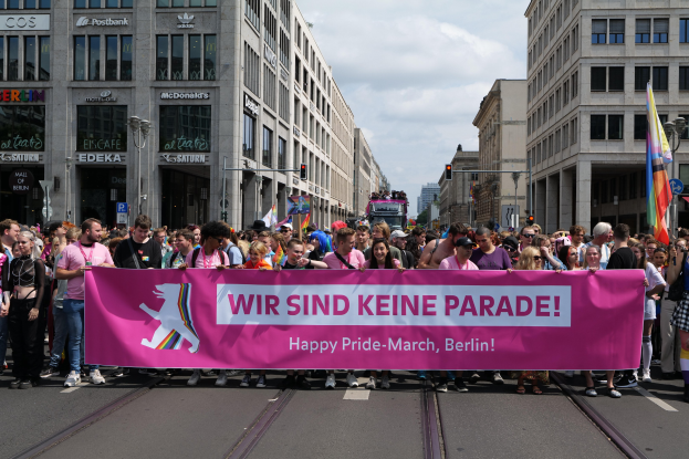 Eine Gruppe von Menschen geht auf einer Straße in Berlin, Deutschland, mit einer pinken Fahne, auf der "Happy Pride March" steht, vorbei an Gebäuden, Laternenmasten und Verkehrszeichen mit einem bewölkten Himmel im Hintergrund.
