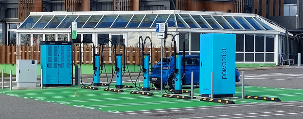 Eine Ladesäule für Elektroautos mit einem blauen Auto in der Mitte, umgeben von blauen und schwarzen Objekten, Pfählen, Brettern und einem Zaun, mit Gebäuden, Bäumen und Himmel im Hintergrund.