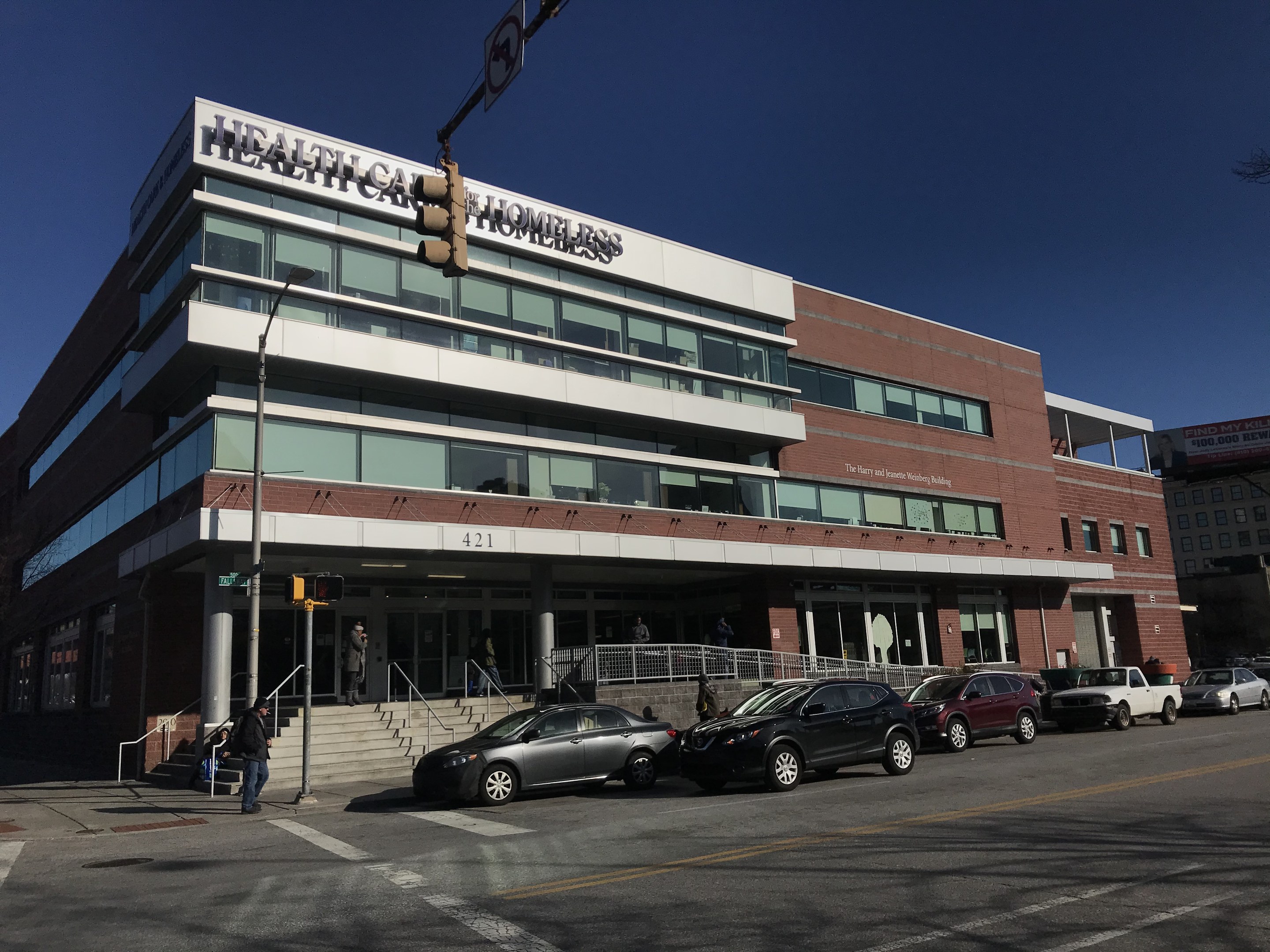 Großes Gebäude mit der Aufschrift "New York City Health Care Homeless Center" mit parkenden Autos, Fußgängern, Straßenmöbeln und Bäumen unter einem klaren Himmel.