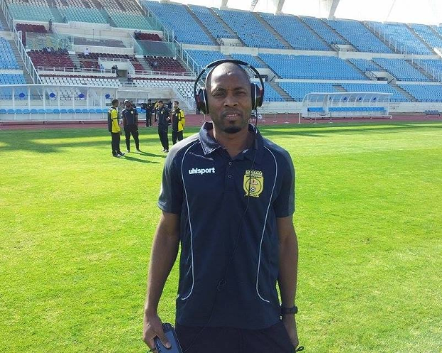 Ein Mann in einem Headset, wahrscheinlich der Trainer der ghanaischen Fußballnationalmannschaft, steht auf einem Fußballfeld neben einem Stadion und hält ein Objekt in der Hand, während im Hintergrund einige Menschen zu sehen sind und der Himmel sichtbar ist.