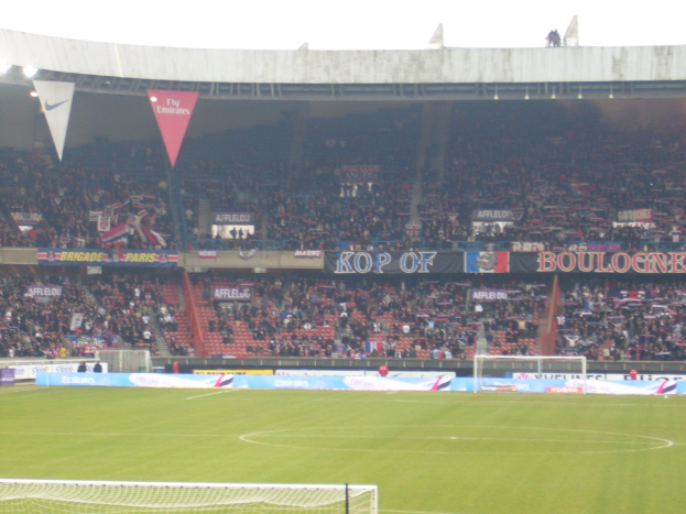Ein Rasenplatz mit Torpfosten auf beiden Seiten, zahlreichen Zuschauern im Hintergrund und vielen Werbetafeln.