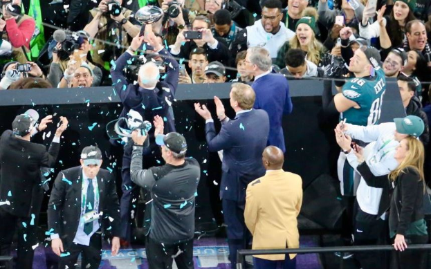 Eine Gruppe von Philadelphia Eagles Spielern feiert mit dem Vince Lombardi Trophy, trägt Kappen und wirft Konfetti, mit einem Geländer und kameraschwingenden Individuen im Hintergrund.