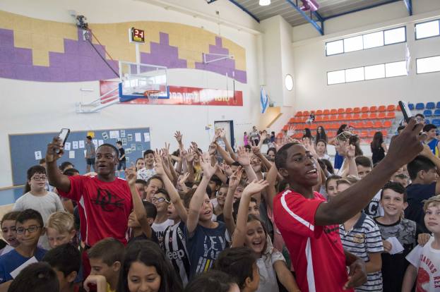 Gruppe von Kindern vor einem Basketballfeld mit Mobiltelefonen in der Hand, mit einer Tafel, einer Uhr, einem Torpfosten, einem Basketballkorb, Deckenleuchten, Stühlen und Fenstern im Hintergrund.