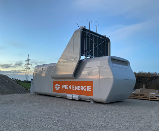 Eine große weiße und orangefarbene Windkraftanlage steht auf einem Schotterfeld umgeben von Bäumen und einem Turm im Hintergrund, mit einigen Gegenständen auf der rechten Seite und dem Himmel oben.