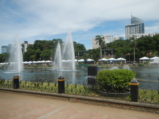 Ein Brunnen in einem Park umgeben von einem Zaun, Pflanzen, Gras, Menschen, Fahrzeugen, Zelten, Fahnen, einem Straßenschild, Bäumen, Gebäuden mit Fenstern unter einem bewölkten Himmel.