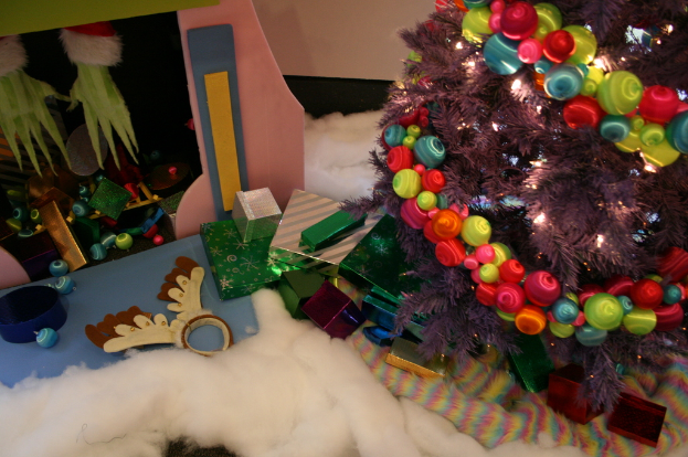 Eine Indoor-Szene mit einem geschmückten Weihnachtsbaum auf der rechten Seite, Geschenkboxen und Dekorationen in der Mitte und einer Hütte mit Dekorationen oben links in der Ecke, mit Watte und Wolle unten.